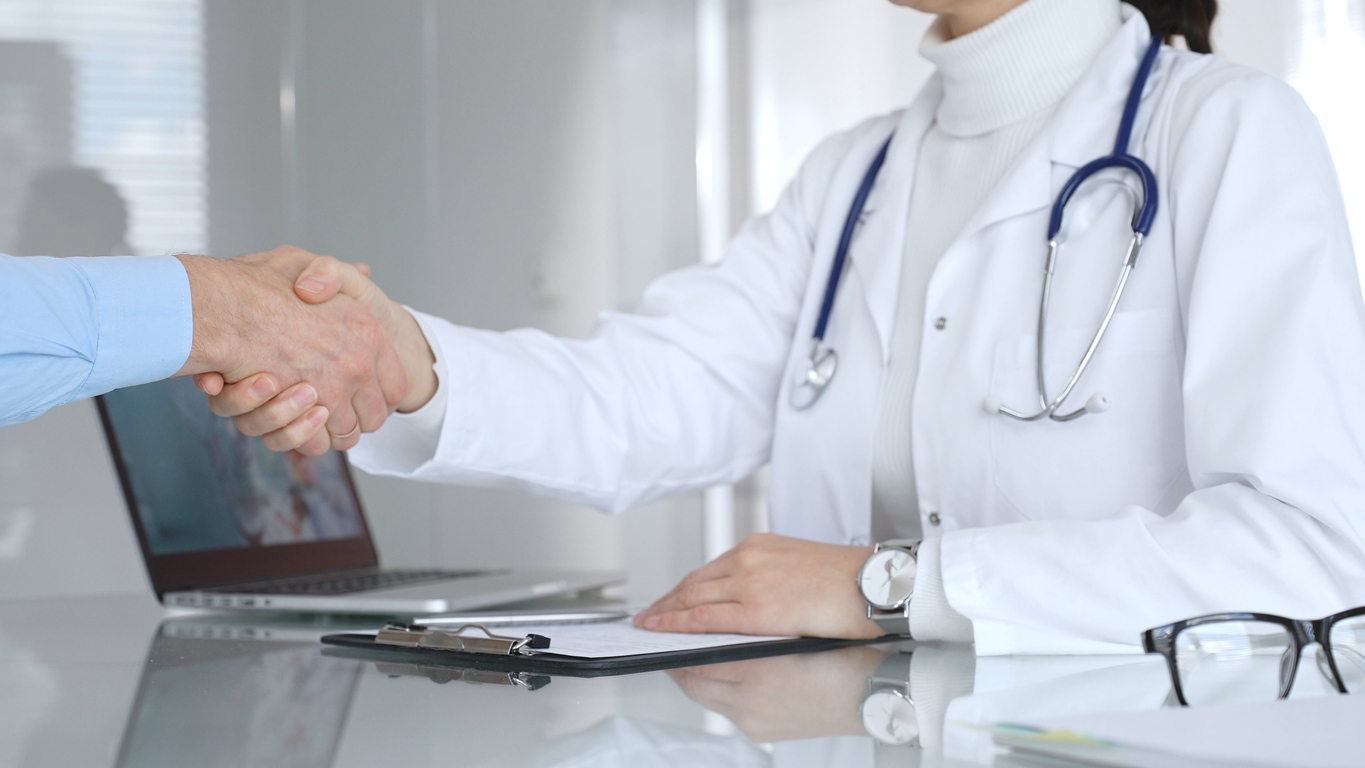 Hands of medical professional and male shaking across a desk, symbolizing trust and partnership.